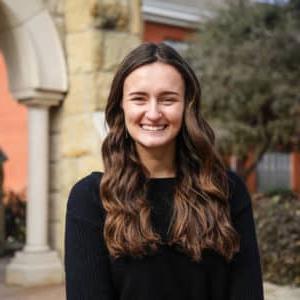 A smiling woman with long wavy hair, wearing a black sweater, standing in front of Howard Payne University's building with stone arches. | HPU